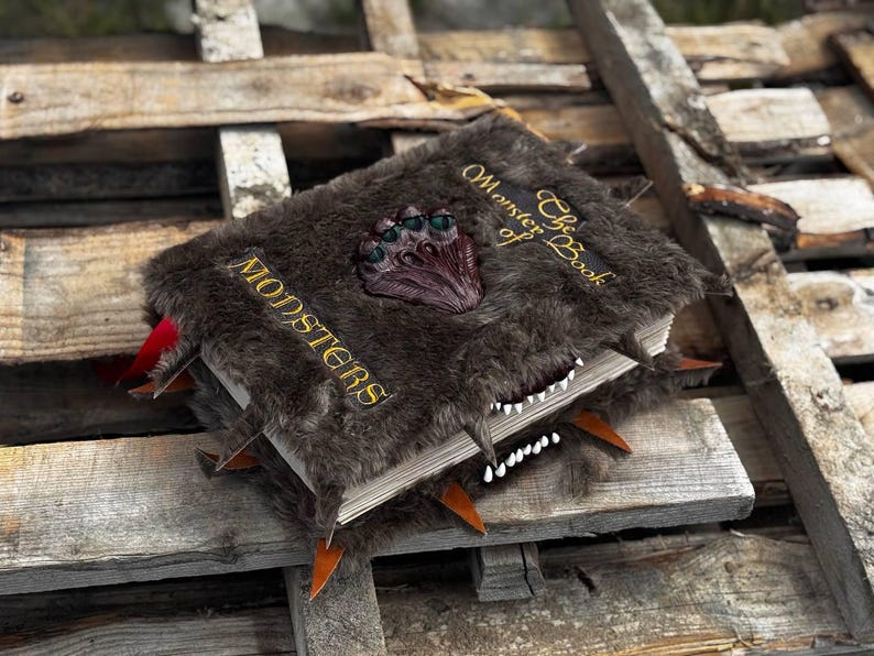 May include: A faux fur-covered book titled "The Monster Book of Monsters" with a monster claw clasp. The book has a toothy grin and orange spikes. The word "MONSTERS" is embroidered in gold.