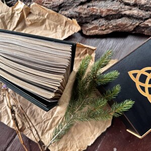 May include: A black book with a gold ribbon bookmark lies open on a brown paper surface. A green pine branch and a black box with a gold Celtic knot design are also visible.