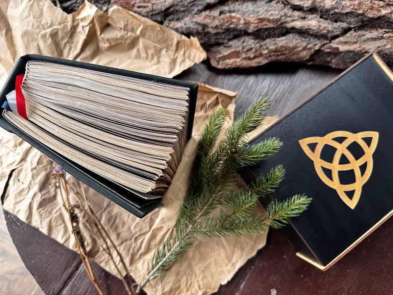 May include: A black book with a gold ribbon bookmark lies open on a brown paper surface. A green pine branch and a black box with a gold Celtic knot design are also visible.