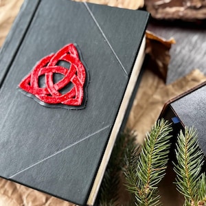 May include: A black leather-bound journal with a red three-dimensional Celtic knot design on the front cover.