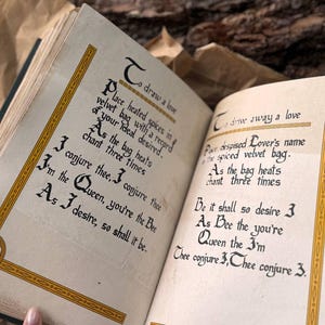 May include: An open book with yellowed pages and gold trim. The text on the pages is in a handwritten font and reads "To draw a love" and "To drive away a love". The text describes a spell using a velvet bag and heated spices.