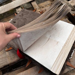 May include: An open, aged book with off-white pages, held by a hand. The book has a worn appearance, with visible aging marks. The pages are thick and textured. The book is resting on a wooden surface with decorative elements.