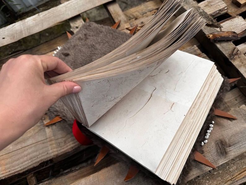 May include: An open, aged book with off-white pages, held by a hand. The book has a worn appearance, with visible aging marks. The pages are thick and textured. The book is resting on a wooden surface with decorative elements.