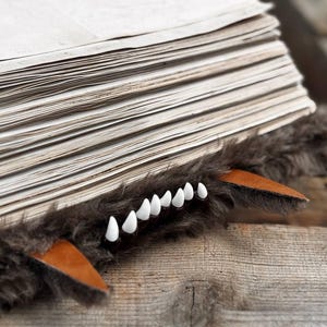 May include: A whimsical book accessory featuring a faux fur wolf design. The accessory has a stack of pages with a brown fur trim, leather accents, and white teeth. The design is set on a wooden surface.