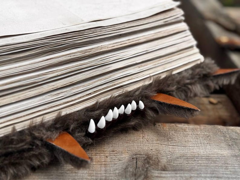 May include: A whimsical book accessory featuring a faux fur wolf design. The accessory has a stack of pages with a brown fur trim, leather accents, and white teeth. The design is set on a wooden surface.