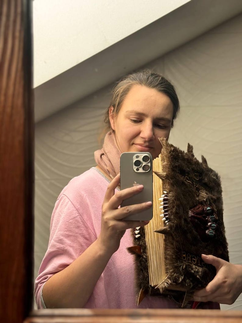 May include: A large, brown, faux-fur covered book with a spine of white teeth and decorative metal accents. The book has a weathered appearance and is held in front of a mirror.