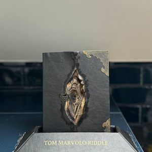 May include: A black leather book with a distinctive tear-shaped design exposing a lighter interior. The corners feature decorative brass details. The base is inscribed with the words "TOM MARVOLO RIDDLE".