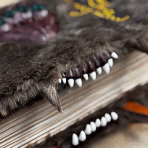May include: A detailed shot of a fantasy book with a dark gray, furry cover. The book features white teeth and a sharp, pointed tooth. The cover has a textured appearance with decorative elements.