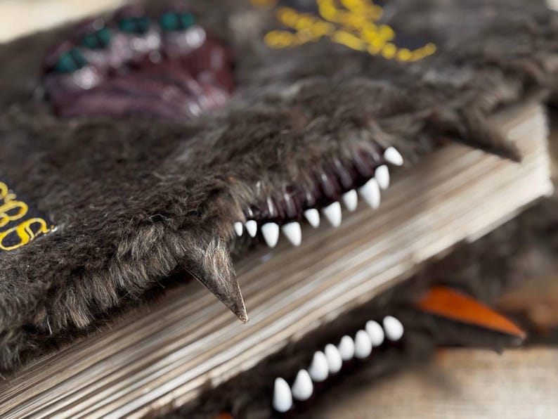 May include: A detailed shot of a fantasy book with a dark gray, furry cover. The book features white teeth and a sharp, pointed tooth. The cover has a textured appearance with decorative elements.
