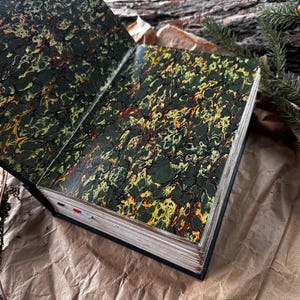 May include: An open book with a marbled green, yellow, and red cover. The book is lying open on a brown paper surface.