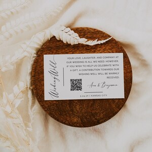 May include: A white card with black text that reads "Wishing Well" in cursive. The card also includes a QR code, the text "Your love, laughter, and company at our wedding is all we need. However, if you wish to help us celebrate with a gift, a contribution towards our wishing well will be warmly received." and the names "Ave & Benjamin". The card also includes the text "5.24.2X | Kansas City".