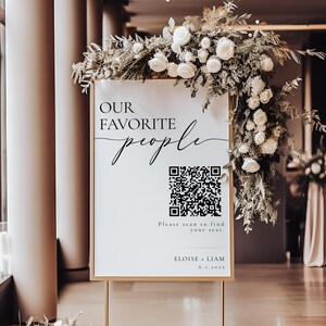 Könnte beinhalten: Ein weißes Schild mit goldenen Verzierungen auf einem goldenen Ständer mit einem schwarz-weißen QR-Code. Das Schild trägt die Aufschrift "Our Favorite People" in einer kursiv geschriebenen Schrift. Unter dem QR-Code befindet sich der Text "Please scan to find your seat." und "Eloise + Liam 6.1.2023". Das Schild ist mit weißen Blumen und Grünpflanzen verziert.