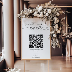 以下が含まれることがあります： 「your seat awaits」の文字とQRコードが書かれた、金色のフレームの看板。白い花と緑で飾られています。看板は金色の金属製のスタンドに立っています。白い花のブーケがスタンドの土台にあります。