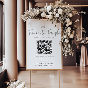以下が含まれることがあります： 黒と白のQRコードがついた金色のスタンドに置かれた、金色の枠のある白い看板。看板には「私たちのお気に入りの人々、あなたの席はあなたを待っています。」と書かれています。QRコードの下には「席を見つけるためにQRコードをスキャンしてください。」というテキストがあります。看板には「Olivia & Hunter」という名前と「9.2.202X」という日付と「カンザスシティ」という場所も記載されています。