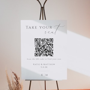 以下が含まれることがあります： 黒文字で「TAKE YOUR seat」と書かれた白い看板で、その下に白黒のQRコードがあります。「Scan the QR code to find your seat」という文字がQRコードの下にあり、その下に「KATIE & MATTHEW 5.4.2X」という文字があります。看板の下部には「KM」のイニシャルがあります。
