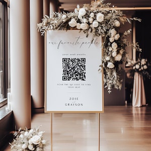 以下が含まれることがあります： 金色の枠のある白い看板が、金色のスタンドの上に置かれています。看板には、白黒のQRコードと「our favorite people」、「your seat awaits」、「SCAN THE QR CODE TO FIND YOUR SEAT」、「ZOIE & GRAYSON」の文字が書かれています。看板は白い花で飾られています。