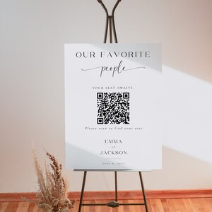 以下が含まれることがあります： 黒いイーゼルに立てられた白い看板。テキスト「OUR FAVORITE people」とQRコードが表示されています。QRコードの下には「Please scan to find your seat」と書かれています。看板には「EMMA and JACKSON」という名前と「June 6, 2025」という日付も含まれています。