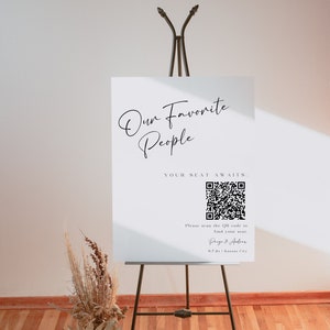 Könnte beinhalten: Ein weißes Schild mit schwarzem Text, der "Unsere Lieblingsmenschen" und "Ihr Platz wartet." Ein QR-Code wird mit dem Text "Bitte scannen Sie den QR-Code, um Ihren Platz zu finden." angezeigt. Das Schild enthält auch die Namen "Paige & Andrew" und den Ort "0.7.2x | Kansas City".