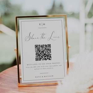 May include: A white sign in a gold frame with the text "Share the Love" and a QR code. The sign also includes the text "WE'D LOVE TO SEE YOUR PHOTOS! Scan the QR code with your smartphone and upload to share with us." and "KATIE & MATTHEW".