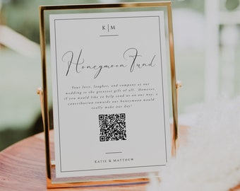May include: A gold-framed sign with a white background and black text that reads "KM Honeymoon Fund". The sign also includes a QR code and the text "Your love, laughter, and company at our wedding is the greatest gift of all. However, if you would like to help send us on our way, a contribution towards our honeymoon would really make our day! Katie & Matthew."