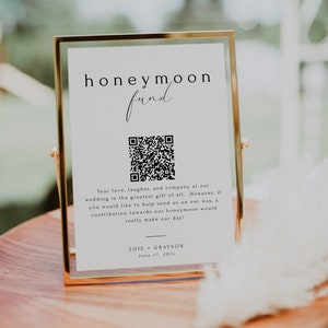 Könnte beinhalten: Ein weißes Schild mit goldenem Rahmen, das einen Hochzeitsreisefonds ankündigt. Das Schild enthält einen QR-Code und den Text "honeymoon fund", "Your love, laughter, and company at our wedding is the greatest gift of all. However, if you would like to help send us on our way, a contribution towards our honeymoon would really make our day!" und "ZOIE + GRAYSON June 17, 202x".