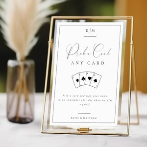 Może przedstawiać: Złota metalowa ramka z napisem "Pick a Card, ANY CARD" w eleganckiej czcionce. Poniżej cztery kolory kart do gry. Znak zawiera nazwiska "KATIE & MATTHEW".