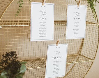 May include: Three white cards with black text on a gold wire rack. The cards are labeled "Table One", "Table Two", and "Table Three" with a list of names under each table number.