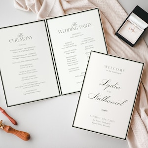 Op de afbeelding: Bruiloft briefpapier set met ceremonie- en bruiloftsgastprogramma's, en een welkomstbord. De programma's hebben een zwarte rand en elegante tekst. Een ringdoosje met een gouden ring, gouden trouwringen en een lakstempel zijn ook aanwezig.
