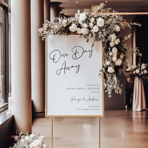 Może przedstawiać: Biały znak z pozłacanym obramowaniem, stojący na złotej podstawce, z zielenią i białymi kwiatami przewieszonymi nad nim. Znak głosi "One Day Away" i jest przeznaczony na kolację próbną celebrującą Paige & Andrew 6 czerwca 202N.