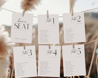 May include: Five white cards with black text, hanging from a string with clothespins. The cards are labeled "Table 1", "Table 2", "Table 3", "Table 4", and "Table 5". Each card has a list of names underneath the table number. The top card says "have a SEAT" with the names "Sophie & Jackson" and the date "July 1, 2023".