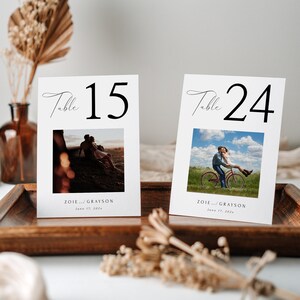 May include: Two white table number cards with black calligraphy. The cards feature a photo of a couple, one on a bicycle and the other sitting on a rock. The text on the cards reads "Table 15" and "Table 24" with the names "ZOIE and GRAYSON" and the date "June 17, 2023".