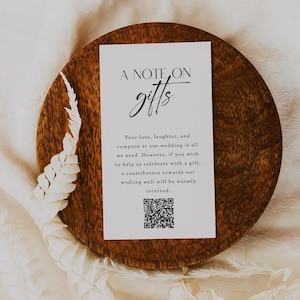 Könnte beinhalten: Eine weiße Karte mit schwarzem Text, der "A Note On Gifts" lautet. Der Text setzt sich fort mit "Deine Liebe, dein Lachen und deine Gesellschaft bei unserer Hochzeit sind alles, was wir brauchen. Wenn du uns jedoch mit einem Geschenk feiern möchtest, ist eine Spende für unseren Wunschbrunnen herzlich willkommen." Ein schwarz-weißer QR-Code befindet sich am unteren Rand der Karte.