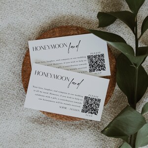 Könnte beinhalten: Zwei weiße Karten mit schwarzem Text und einem schwarz-weißen QR-Code. Der Text auf den Karten lautet "Flitterwochen-Fonds" und enthält eine Nachricht über Hochzeitsgeschenke. Die Karten liegen auf einer Holzoberfläche.
