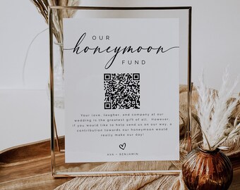 Puede incluir: Un cartel blanco en un marco dorado con un código QR en blanco y negro y el texto "OUR honeymoon FUND". El texto debajo del código QR dice "Your love, laughter, and company at our wedding is the greatest gift of all. However, if you would like to help send us on our way, a contribution towards our honeymoon would really make our day! AVA BENJAMIN".