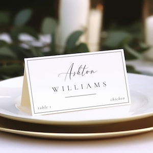 May include: White place card with black border and text. The text reads "Ashton Williams" with a line under the last name. The card also says "table 1" and "chicken".