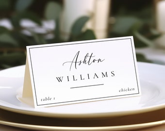May include: White place card with black border and text. The text reads "Ashton Williams" with a line under the last name. The card also says "table 1" and "chicken".