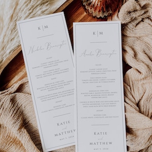 May include: Two white cards with black text outlining the wedding menu for Katie and Matthew. The menu includes salads, main courses, and desserts. The cards are on a brown wooden surface with a beige fabric background.