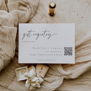 Könnte beinhalten: Eine weiße Karte mit dem Text "gift registry" in schwarzer Kursivschrift. Unter dem Text steht "Madelyn + Sawyer are registered at Target | Amazon | Crate & Barrel" und ein QR-Code ist enthalten. Der Text "www.ourwedding.com" befindet sich am unteren Rand der Karte.