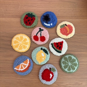 May include: A collection of ten colorful, round, fruit-themed coasters. Each coaster features a different fruit design, including a strawberry, blueberry, cherry, peach, lemon, orange slice, watermelon slice, apple, and lime. The coasters are made with a textured, looped yarn.