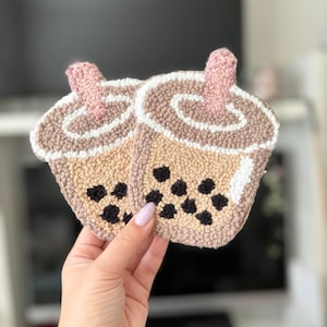 May include: Two brown and beige crocheted bubble tea cups with black boba pearls. The cups have white outlines and pink straws.