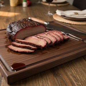 Puede incluir: Una tabla de cortar de madera con una pechuga, parcialmente cortada, y un cuchillo para trinchar. La pechuga tiene un exterior oscuro y crujiente y un interior rosado. La tabla está sobre una mesa de madera.