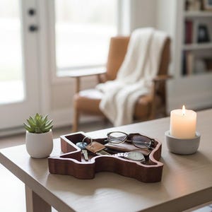 Puede incluir: Una bandeja de madera con forma de Texas contiene llaves, gafas y un reloj sobre una mesa de madera clara. Una pequeña suculenta en una maceta blanca y una vela encendida en un soporte de hormigón también están sobre la mesa. Un sillón de cuero y una ventana están en el fondo.