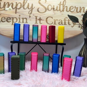 May include: A collection of colorful, rectangular containers with ridged sides, arranged on a black metal shelf and a pink surface. The containers are in various colors, including blue, purple, pink, red, and yellow. The wooden sign in the background reads "Simply Southern Keepin' It Crafty."
