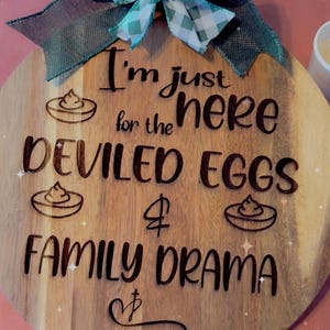 May include: Wooden serving board with a handle, featuring the engraved text "I'm just here for the deviled eggs & family drama." A teal and white checkered bow and green ribbon adorn the top. The board has a round shape.