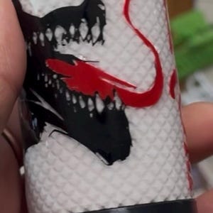 May include: A cylindrical white tumbler featuring a textured diamond-patterned surface. The design displays a black graphic of a creature with sharp teeth and a prominent red tongue. A solid black horizontal band encircles the lower section of the item.