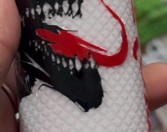 May include: A cylindrical white tumbler featuring a textured diamond-patterned surface. The design displays a black graphic of a creature with sharp teeth and a prominent red tongue. A solid black horizontal band encircles the lower section of the item.