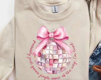 May include: Sand-colored sweatshirt featuring a pink disco ball design with a pink bow and the text "probably just gonna jingle some of the way, I'm tired." The sweatshirt is folded, with a pair of blue jeans and a brown mug visible.