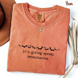 May include: A burnt orange crew neck t-shirt featuring a black graphic of a row of flying bats above the text "it's giving spooky veterinarian" in a typewriter font. The shirt has a Comfort Colors brand label on the inside neck collar.