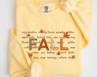 May include: A light yellow sweatshirt with the word "FALL" in brown and orange block letters. The shirt features a list of fall-themed words and phrases in black, with small leaf illustrations.