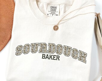 May include: Ivory-colored t-shirt with the words "SOURDOUGH BAKER" in a camouflage-style font. A gold necklace with a pendant rests on the shirt. The shirt is folded and has a Comfort Colors tag.